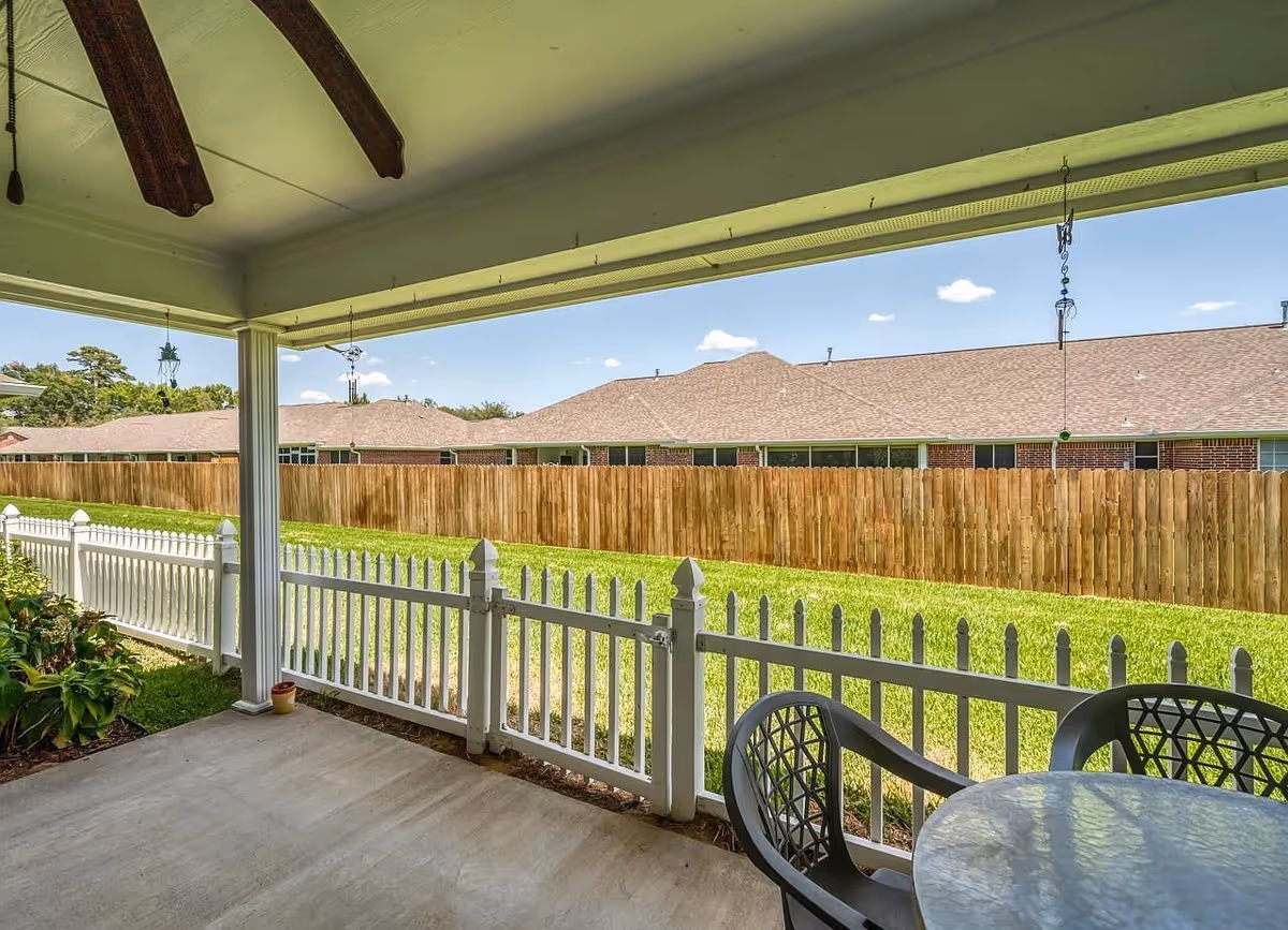 Covered patio area with a ceiling fan, white picket fence, green lawn, and wooden privacy fence. There is a round glass table with black chairs on the patio, and hanging decorations are visible from the ceiling. In the background, there are single-story brick buildings with shingled roofs under a blue sky with a few clouds.
