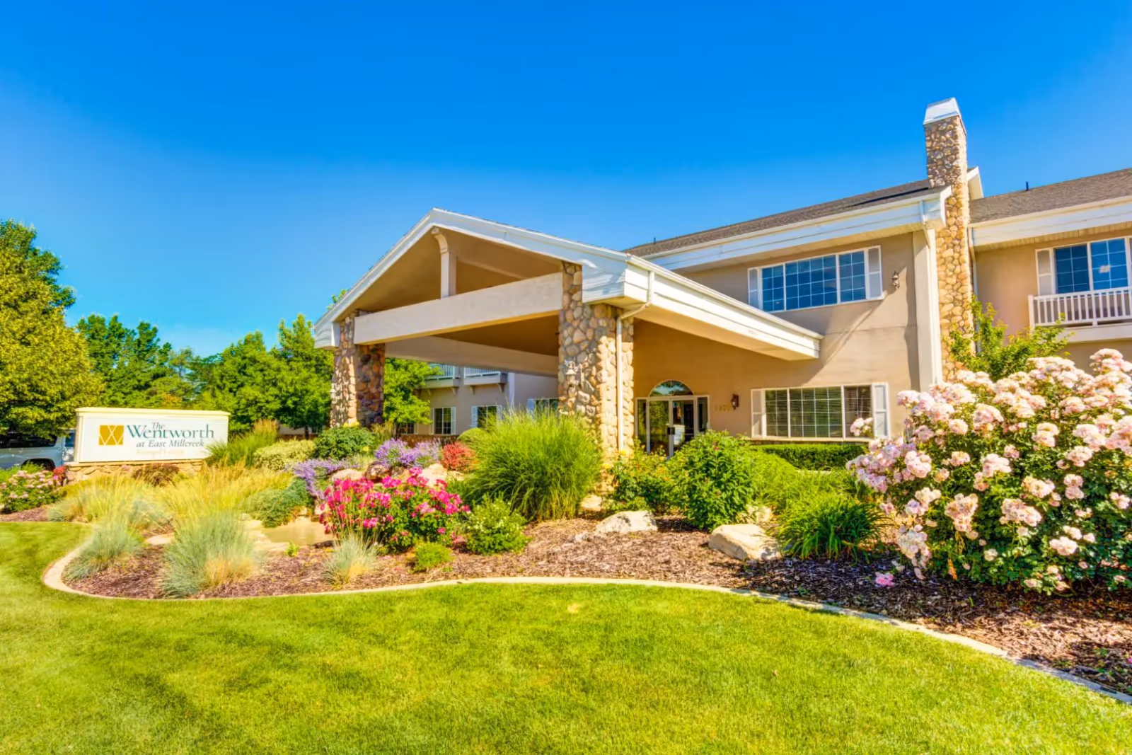 Exterior view of The Wentworth at East Millcreek facility showing a building entrance with stone pillars and a covered driveway. The surrounding area features well-maintained landscaping with green grass, colorful flowers, bushes, and trees under a clear blue sky.