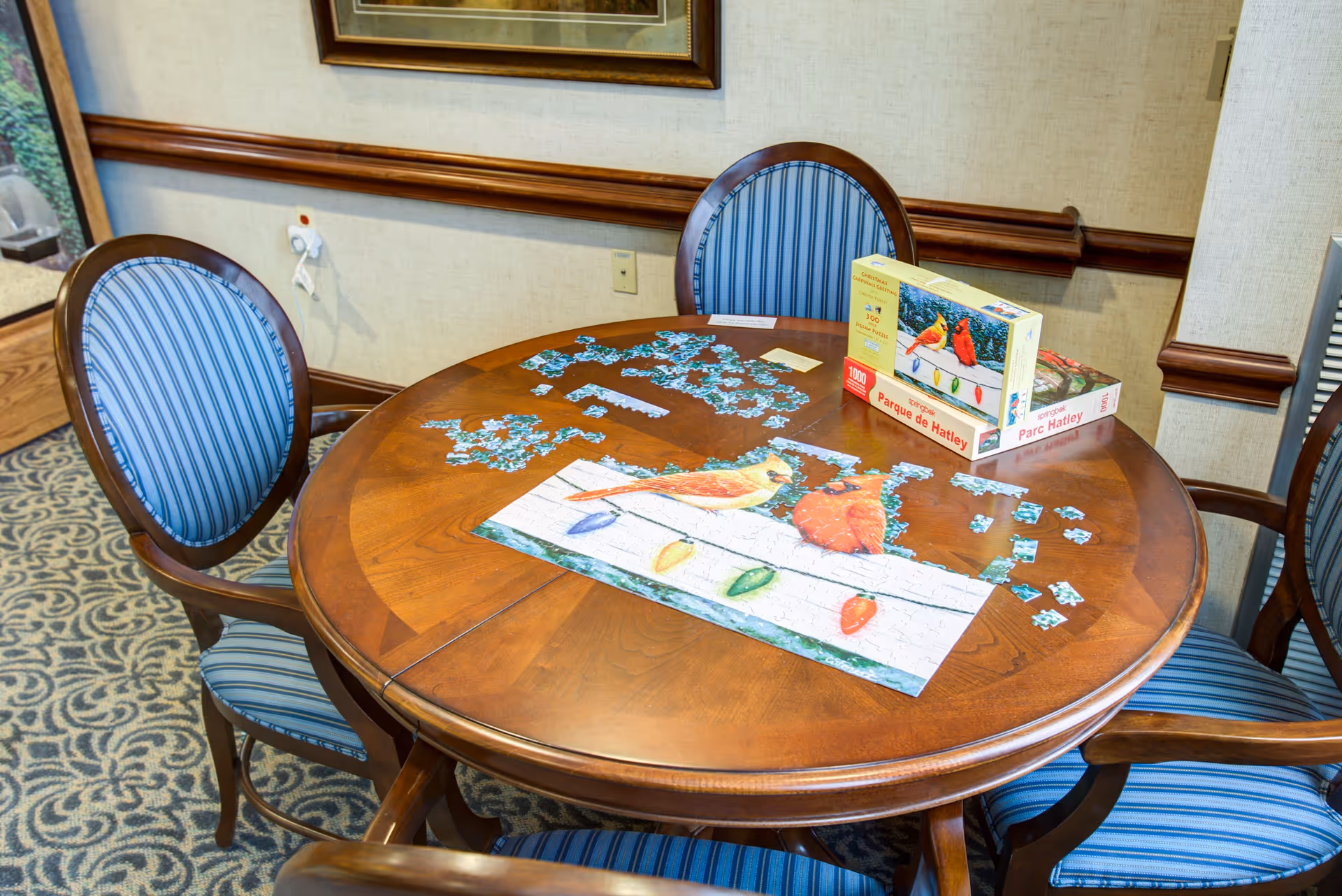 A round wooden table with a partially completed jigsaw puzzle depicting birds and colorful lights. The puzzle box is on the table, and four wooden chairs with blue striped cushions surround the table. The room has patterned carpet and beige walls with wooden trim.