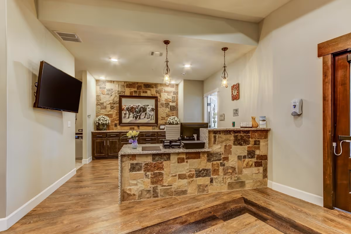 Reception area of Clear Fork Assisted Living and Memory Care featuring a stone front desk with a computer and office supplies, wooden flooring, a wall-mounted TV on the left, and a stone accent wall with a framed picture of horses behind the desk. There are pendant lights hanging from the ceiling and a hand sanitizer dispenser mounted on the right wall near a wooden door.