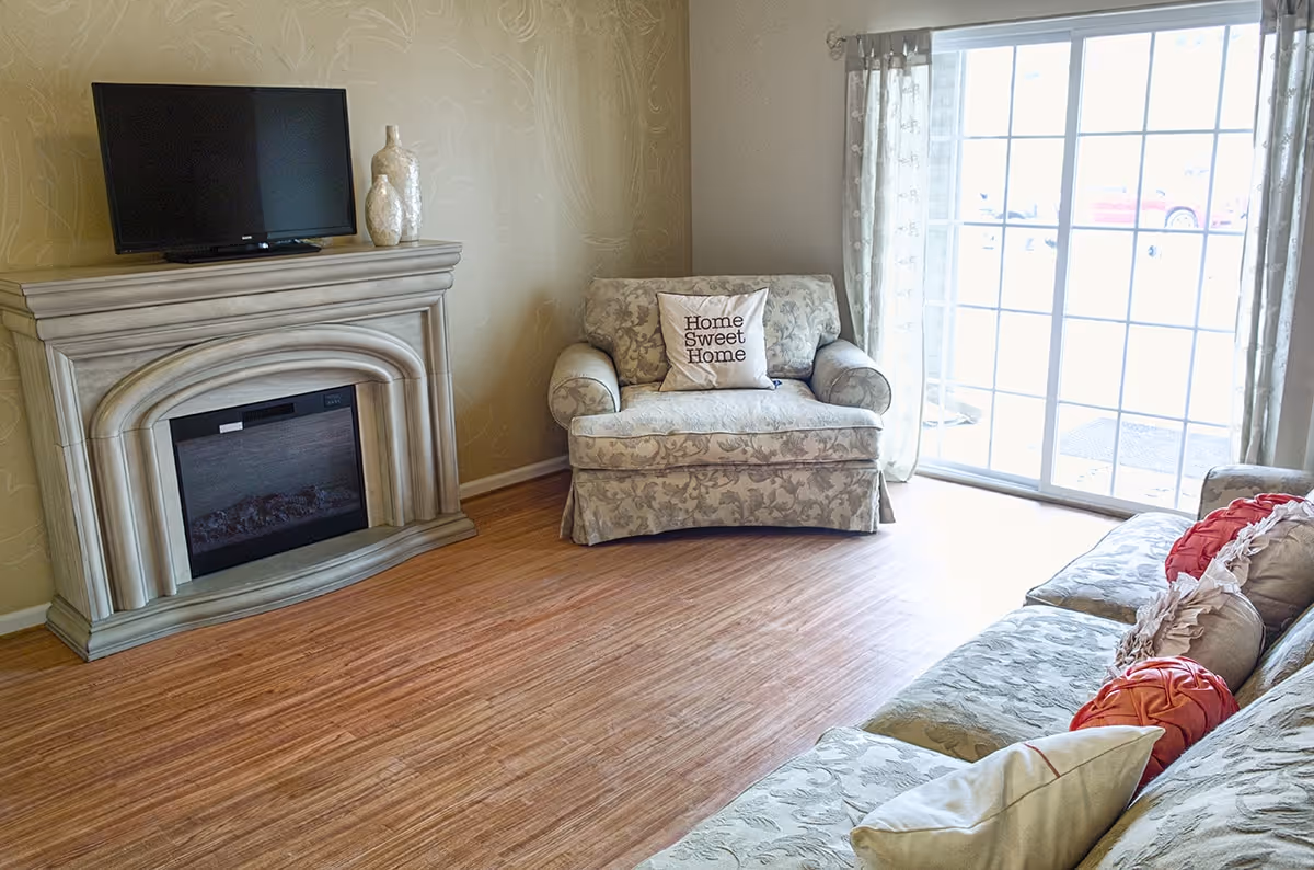 Cozy living room with patterned sofa and armchair (pillow reading "Home Sweet Home"), decorative fireplace with a TV, and sliding glass doors.