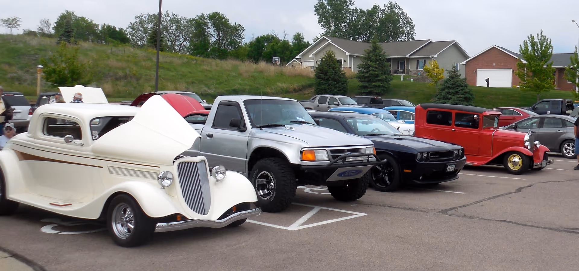 A row of classic and modern cars parked in an outdoor lot with houses and a grassy hill behind them.