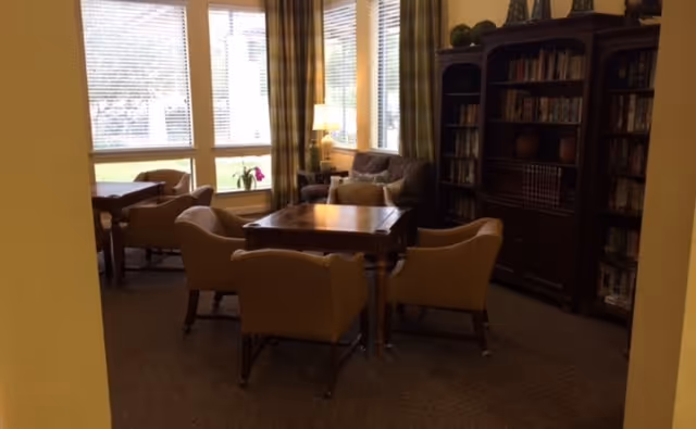 A cozy interior room with a wooden table surrounded by four beige armchairs. Behind the table is a brown sofa with cushions, a floor lamp, and large windows with blinds letting in natural light. To the right, there is a tall wooden bookshelf filled with books and decorative items.