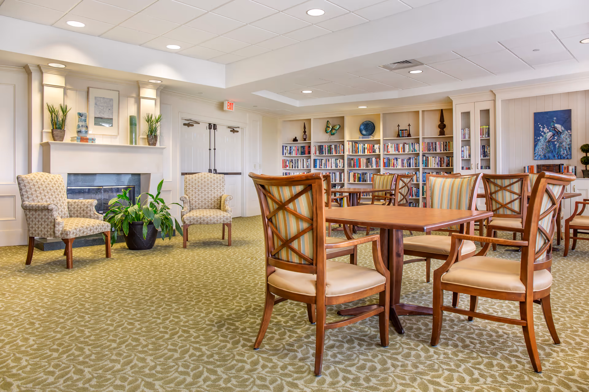A bright and spacious common area with patterned carpet, wooden tables and chairs with cushioned seats, two upholstered armchairs near a fireplace, potted plants, and built-in bookshelves filled with books and decorative items. The walls are light-colored with artwork and the ceiling has recessed lighting.
