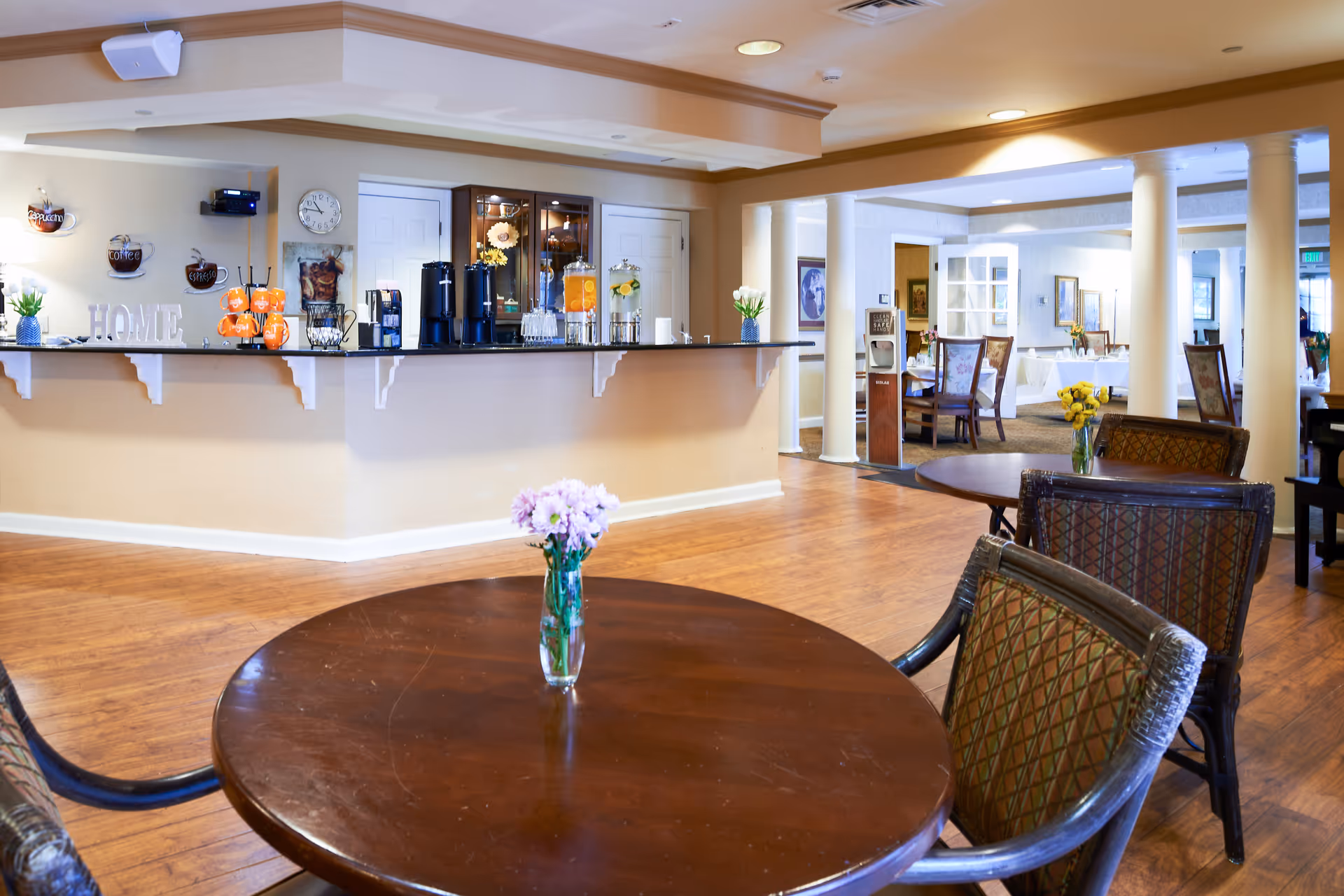 Bright common dining area with round tables and chairs, a serving counter with beverages, and floral centerpieces.