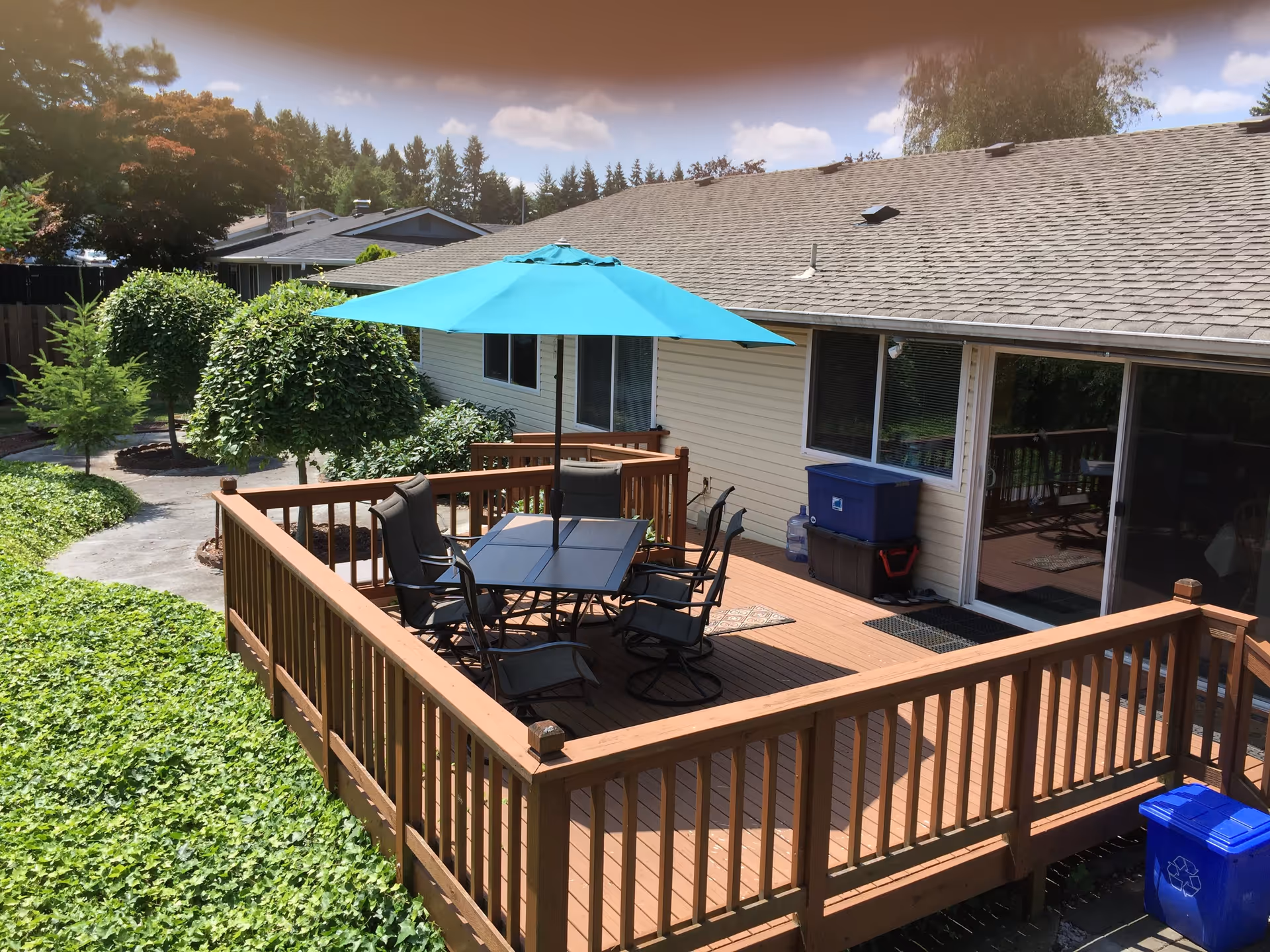 Outdoor wooden deck attached to a beige building with a table and six chairs under a large blue patio umbrella. The deck is surrounded by a wooden railing and overlooks a garden area with green plants and neatly trimmed trees. A blue recycling bin is visible near the deck.