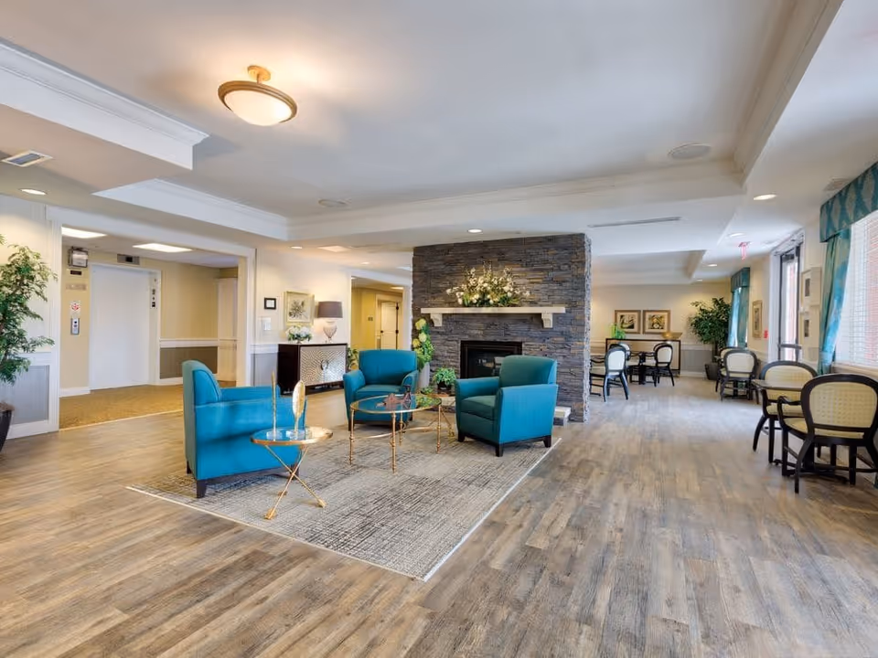 A spacious and well-lit common area with teal armchairs arranged around a glass coffee table on a rug, a stone fireplace with floral decoration on the mantel, and several tables with chairs near large windows with teal curtains. The room has wood flooring and light-colored walls, with plants and framed artwork adding to the decor.