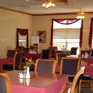 Dining room with multiple tables covered in burgundy tablecloths and wooden chairs beneath ceiling fans and windows.