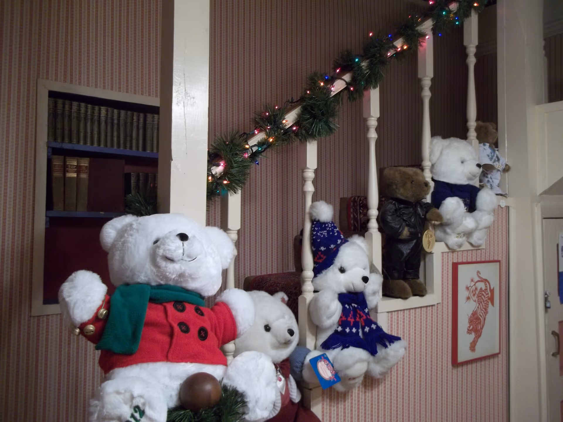A staircase railing decorated with garland and colorful Christmas lights, with several teddy bears dressed in various outfits sitting on the railing spindles. The wall behind has striped wallpaper and a framed picture of a red tiger.