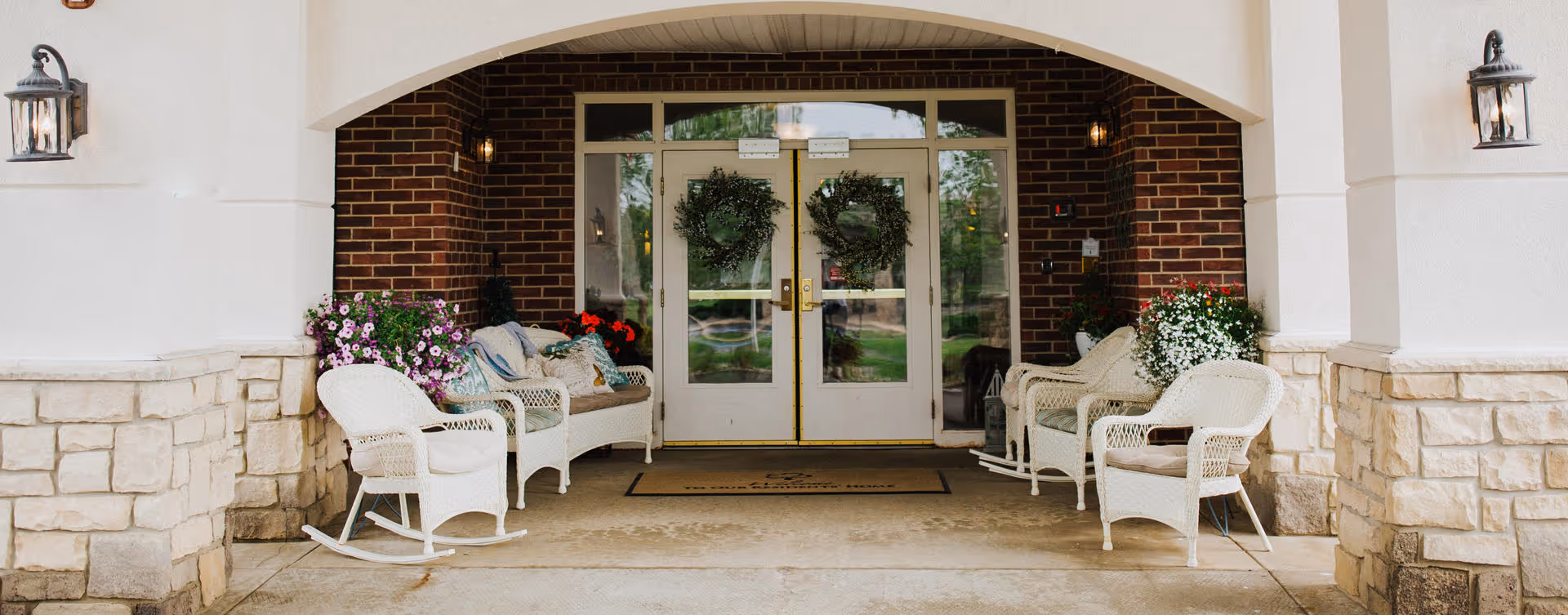 Covered entrance to a building with double glass doors decorated with wreaths. On either side of the entrance are white wicker rocking chairs and benches with cushions, along with potted flowering plants. The walls are a combination of brick and stone with mounted lantern-style lights.