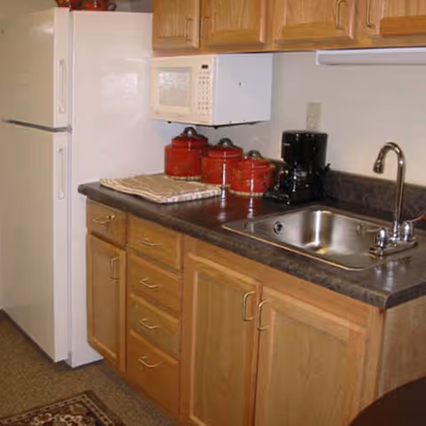 A kitchen area with wooden cabinets, a dark countertop, a stainless steel sink with a faucet, a white microwave mounted above the counter, a white refrigerator, a coffee maker, and three red canisters on the countertop.