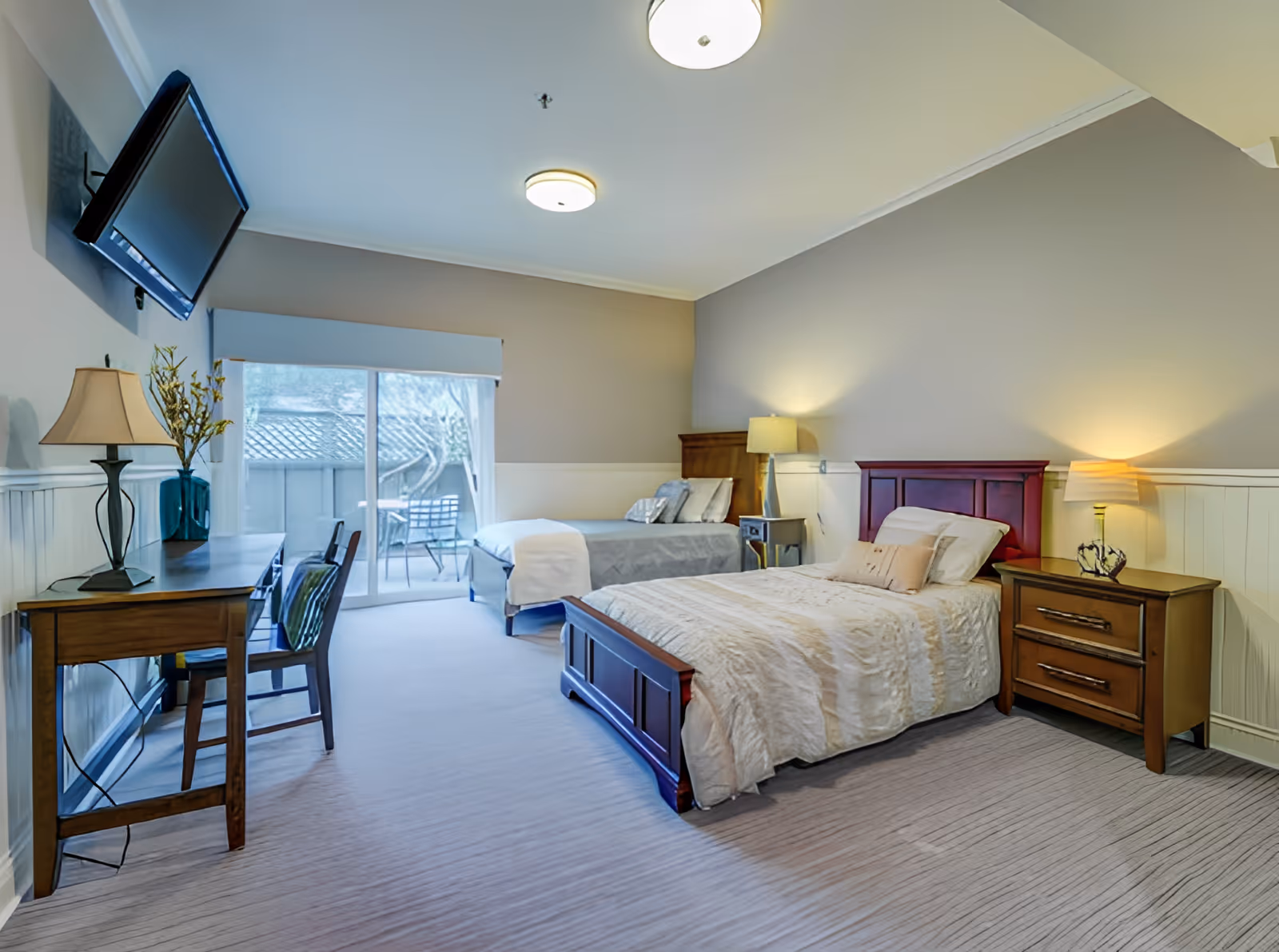 A bright and spacious bedroom with two beds, one with a wooden headboard and beige bedding, the other with a gray bedspread. There are two bedside tables with lamps, a wooden desk with a chair, a flat-screen TV mounted on the wall, and a sliding glass door leading to an outdoor patio with chairs and a table.