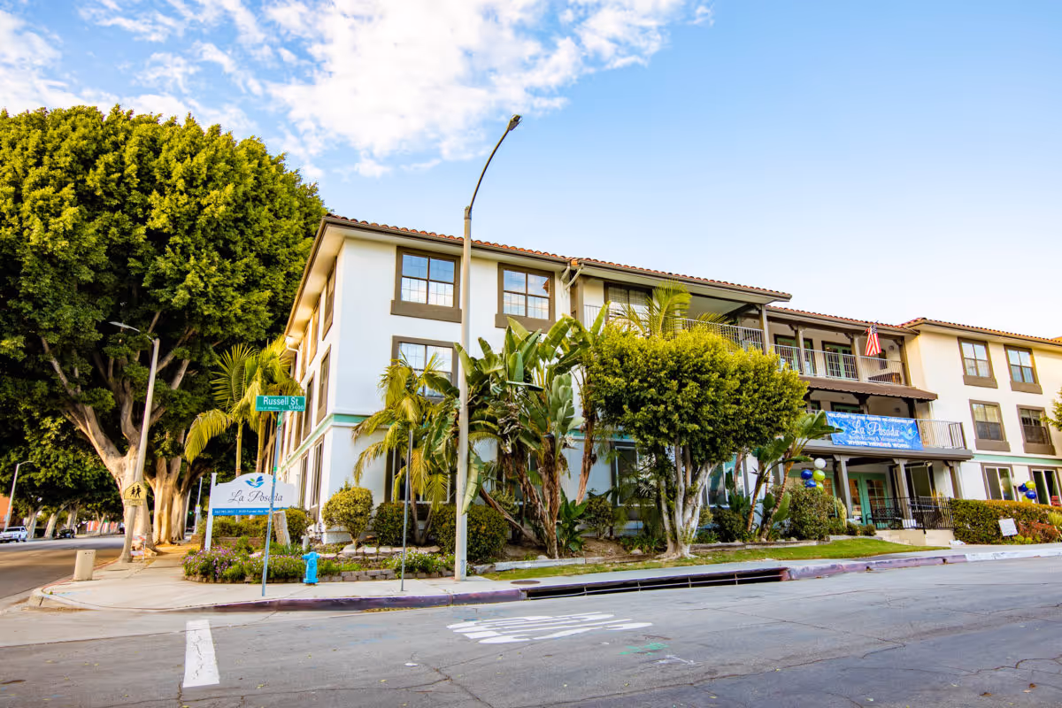Exterior view of La Posada Assisted Living & Memory Care Community building on a sunny day, showing a three-story structure with balconies, surrounded by trees and greenery at the corner of Russell Street.