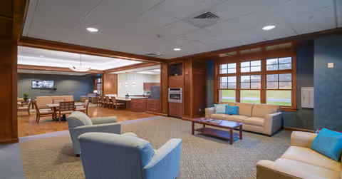 Spacious memory care common room with sofas, armchairs, a coffee table, dining area and large windows.