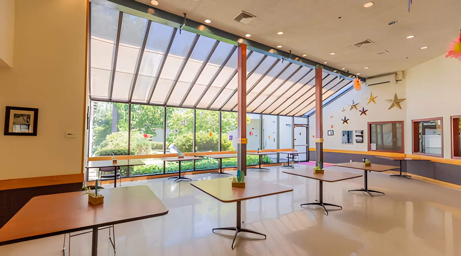 Bright communal dining/activity room with large slanted skylight windows, several spaced tables, and a garden view.