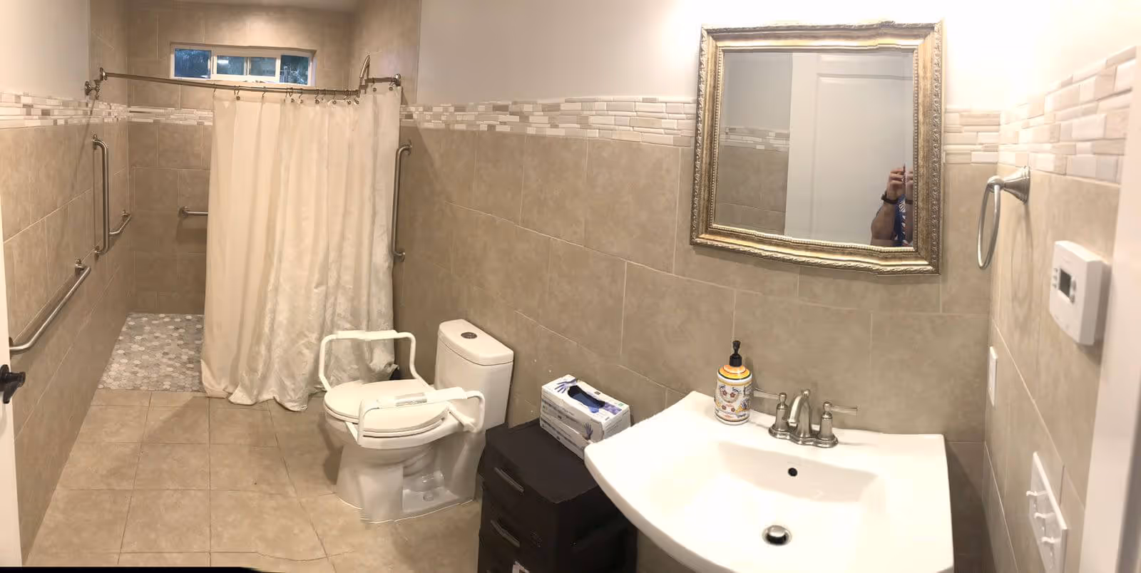 A bathroom with beige tiled walls and floor, featuring a toilet with white safety rails, a walk-in shower with a white curtain, a white sink with a soap dispenser, a decorative framed mirror above the sink, and grab bars along the walls for support.