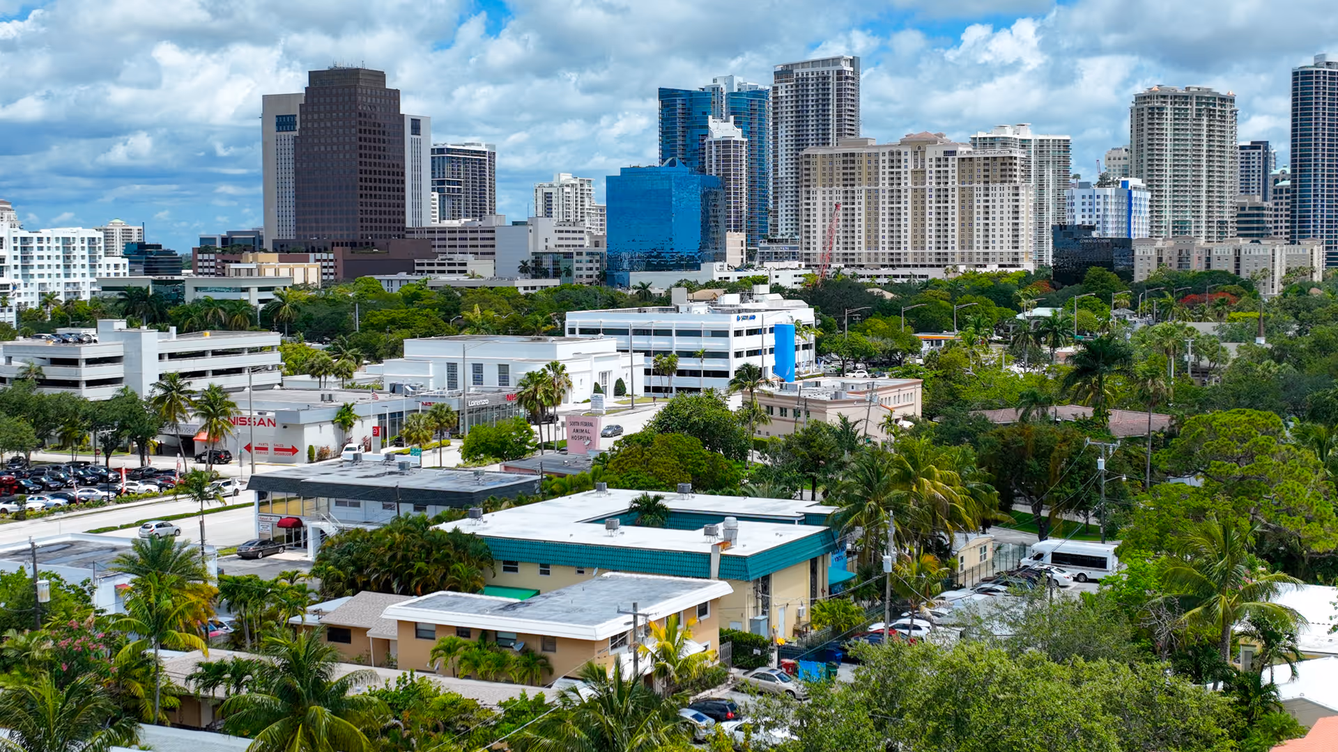 A cityscape view showing a mix of mid-rise and high-rise buildings in the background with a variety of trees and smaller buildings in the foreground under a partly cloudy sky.