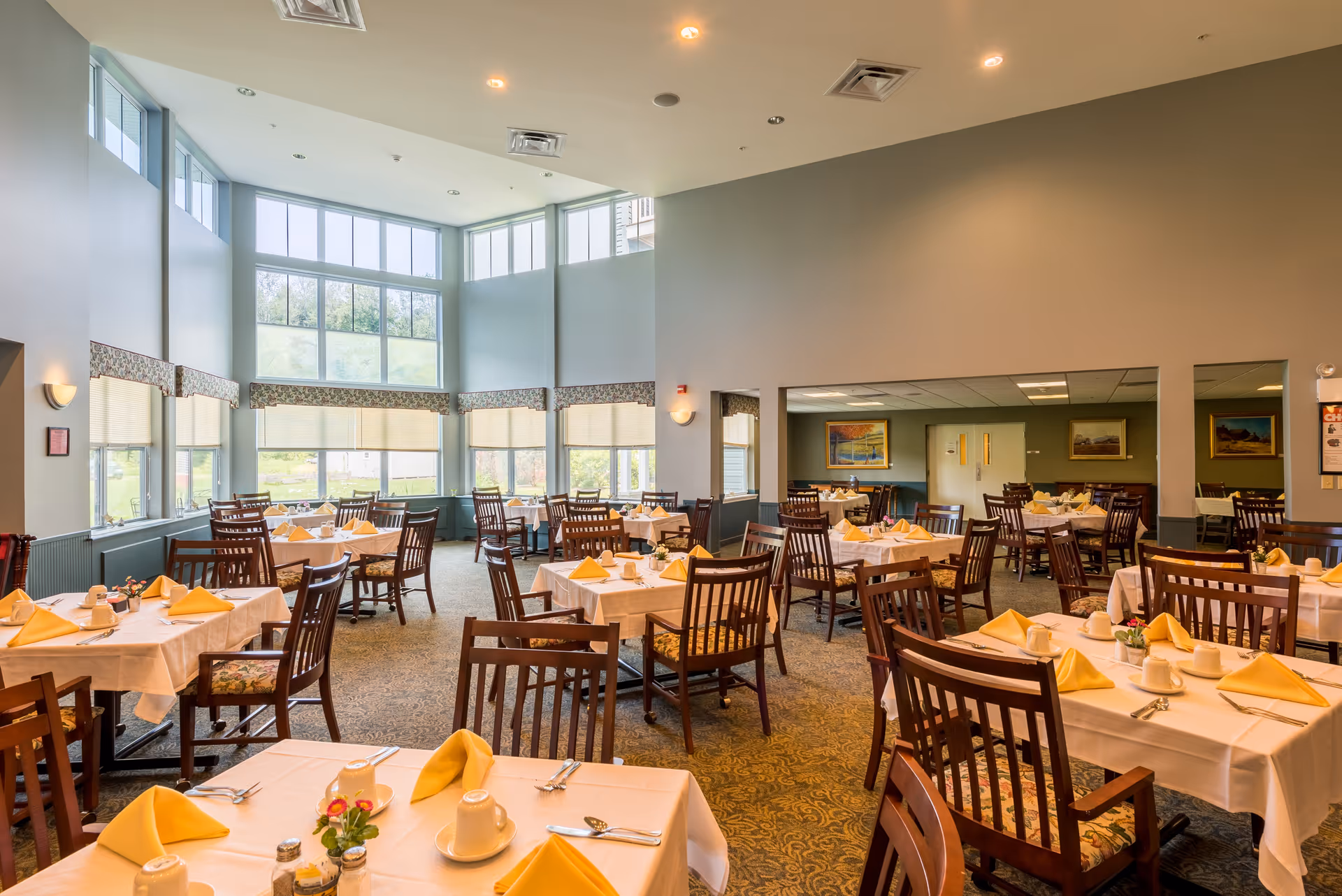 A spacious dining room with multiple tables covered in white tablecloths, each set with yellow folded napkins, white cups, and silverware. The room features large windows allowing natural light to fill the space, wooden chairs with patterned cushions, and soft lighting from ceiling fixtures.