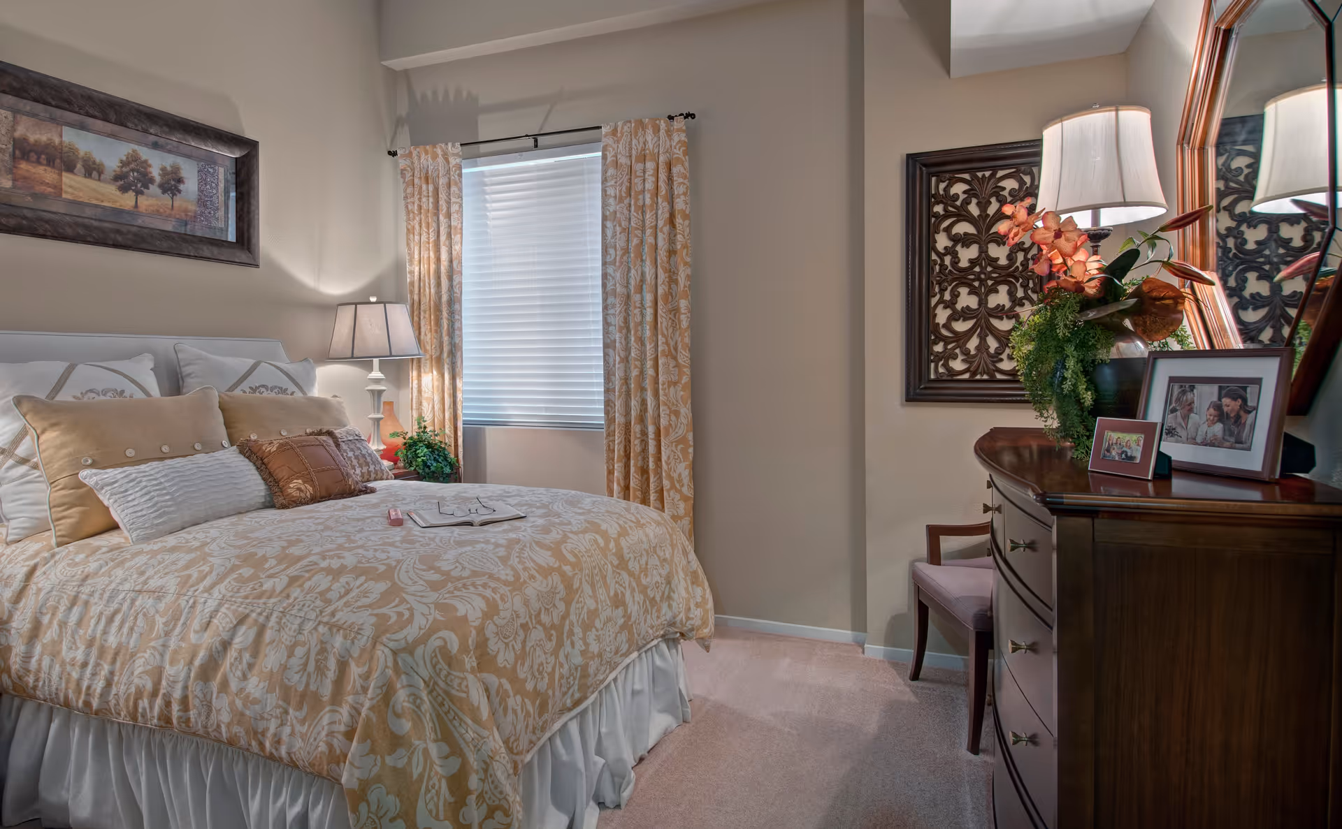 A cozy bedroom with a neatly made bed featuring beige and white patterned bedding and multiple pillows. A bedside table with a lamp and a small plant is next to the bed. A window with beige patterned curtains and blinds is behind the bed. On the right side, there is a wooden dresser with a lamp, a decorative floral arrangement, framed photos, and a mirror above it. A chair is partially visible next to the dresser.
