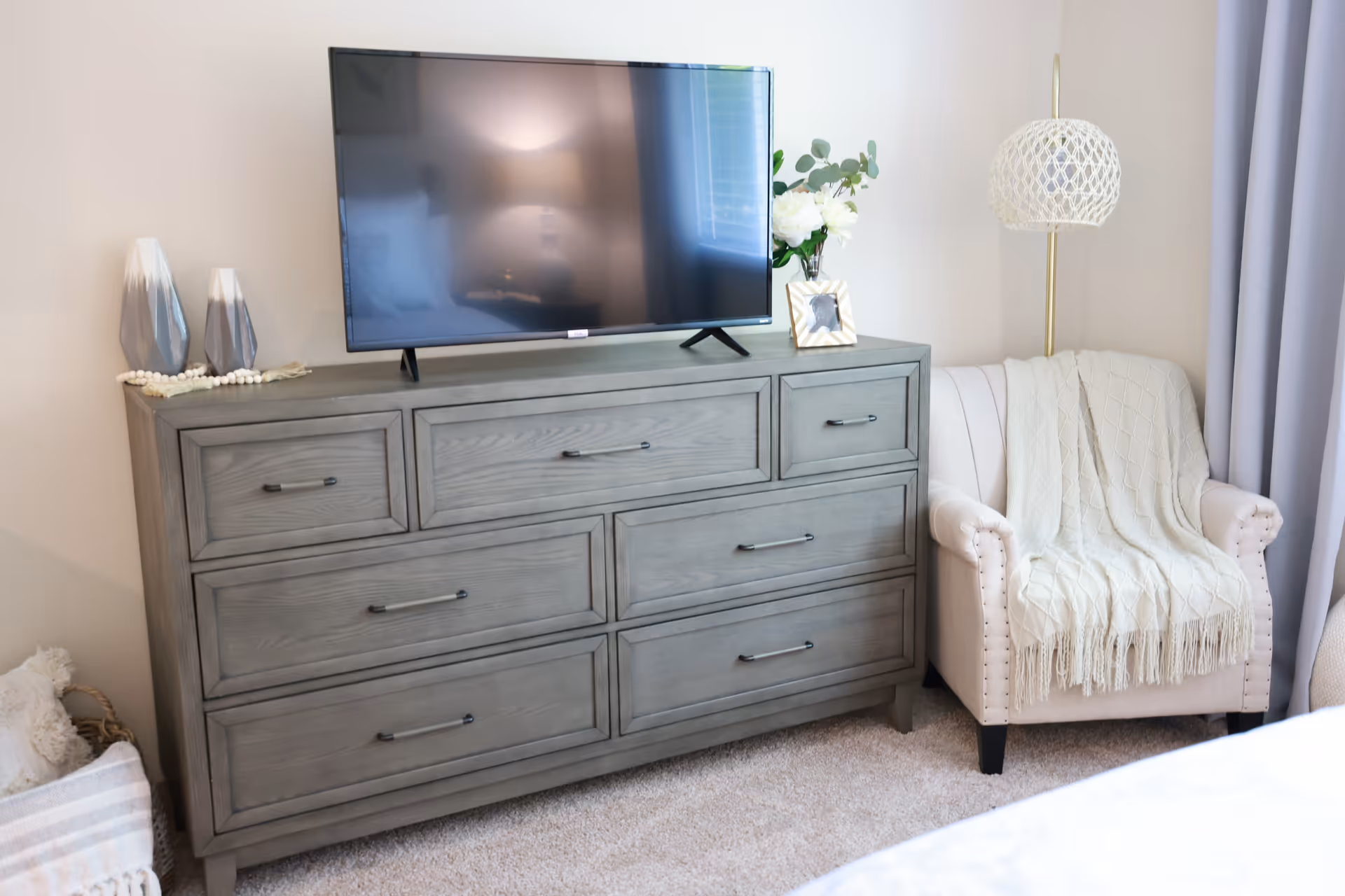 A cozy corner of a bedroom featuring a gray wooden dresser with a flat-screen TV on top, decorative vases, a framed photo, and a vase with white flowers. Next to the dresser is a beige armchair draped with a cream-colored knitted throw blanket, and a floor lamp with a woven lampshade stands behind the chair.