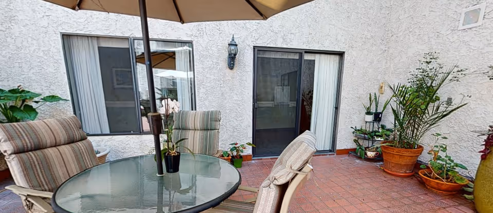Outdoor patio area with a round glass table and four cushioned chairs under a large umbrella. The patio is surrounded by a white textured wall with two windows and a sliding glass door. Several potted plants are arranged along the edges of the patio.