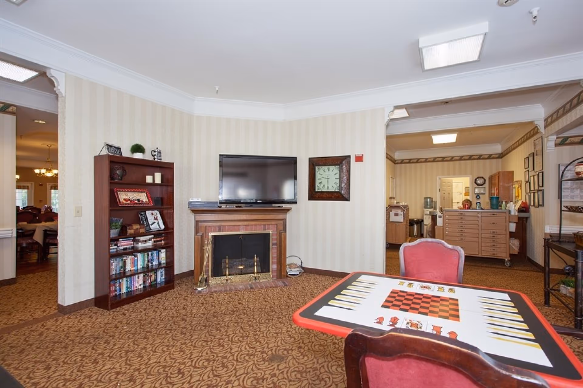 A cozy common room with a TV over a fireplace, a bookshelf, and a game table with chairs.