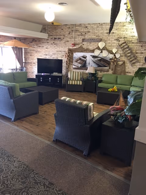 A cozy living room area with green cushioned sofas and dark wicker chairs arranged around two black coffee tables. A flat-screen TV is placed on a black stand against a brick wall. The wall is decorated with a large picture of boats on water, a wooden ladder, and other rustic decor items. The floor is wooden, and there is a patterned carpet leading into the room.