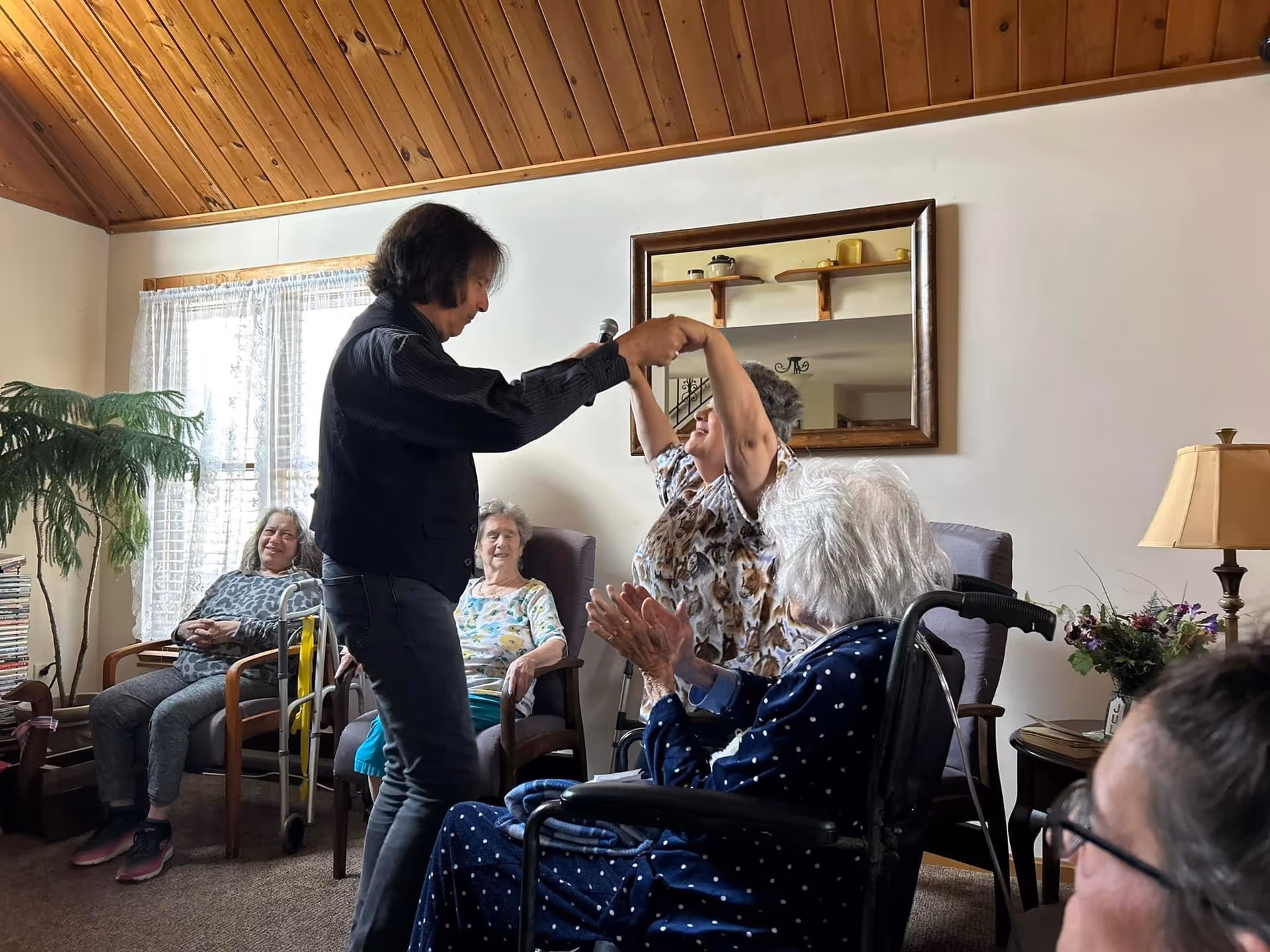 A group of elderly women in a cozy living room setting. One woman is standing and holding hands with another woman who is seated, appearing to dance or engage in an activity together. Other women are seated around them, some in wheelchairs and others in chairs, watching and smiling. The room has a wooden ceiling, a large window with sheer curtains, a potted plant, a lamp, and a mirror on the wall reflecting part of the room.
