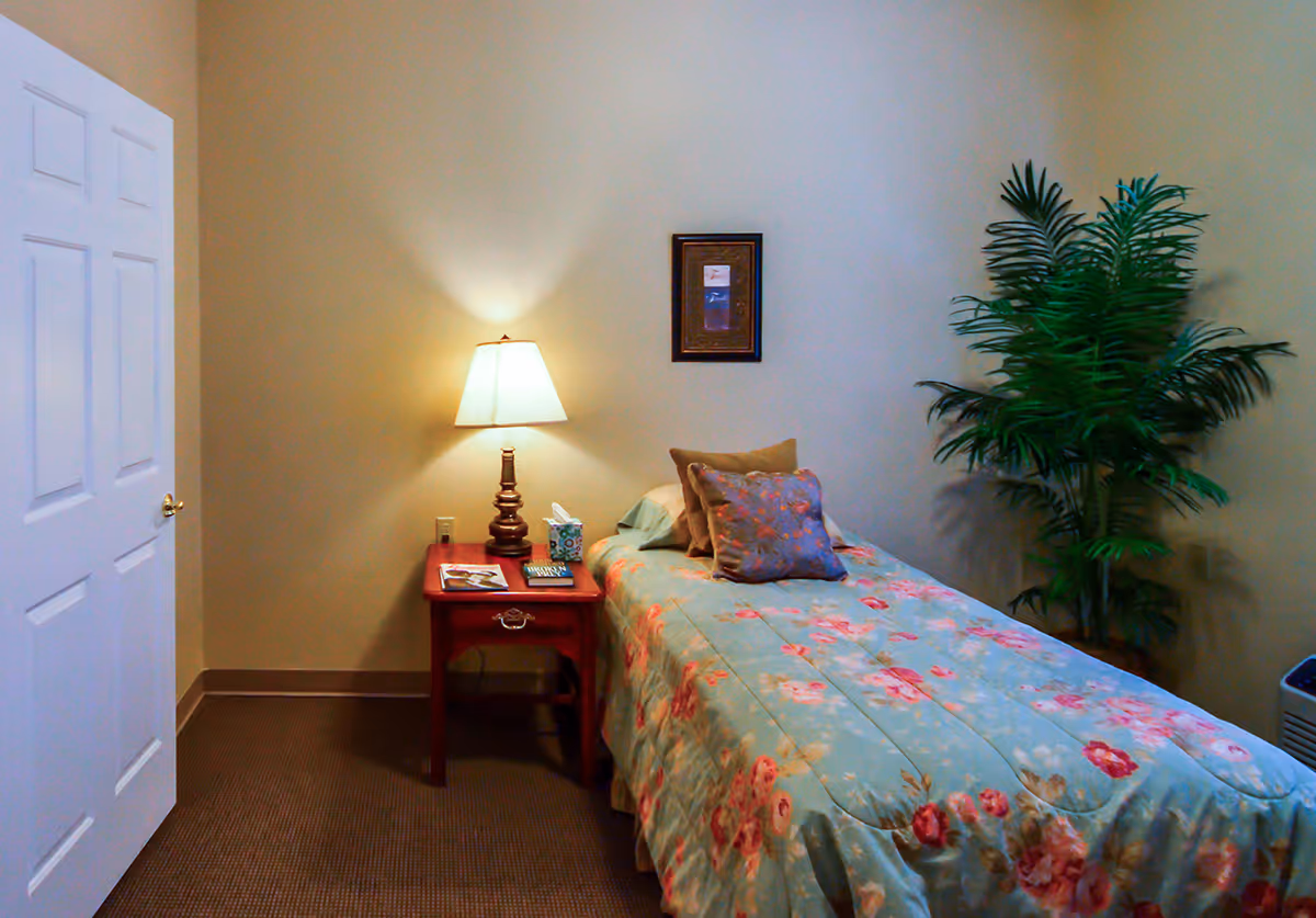 A small bedroom with a single bed covered in a floral bedspread. There are two pillows on the bed, one beige and one with a floral pattern. Next to the bed is a wooden nightstand with a lamp, a tissue box, and some books. A framed picture hangs on the wall above the bed, and a large green potted plant is placed in the corner of the room. The walls are painted beige, and there is a white door on the left side.