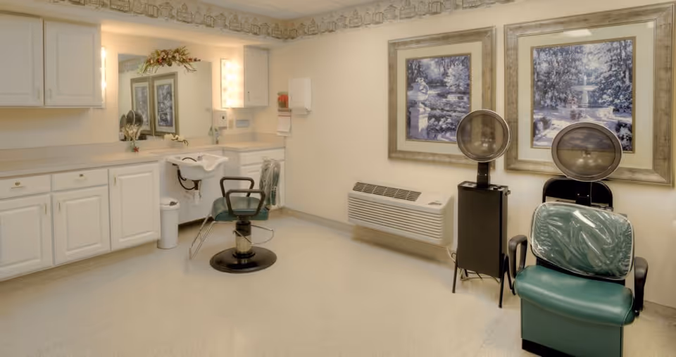 Interior of a salon area in a senior living facility with two green salon chairs, a hair washing sink, white cabinets, two framed pictures on the wall, and hair drying machines.