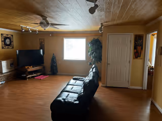 A living room with wooden flooring and a wooden ceiling. There is a black leather couch facing a flat-screen TV on a wooden stand. The room has a window with bright light coming through, a ceiling fan, and some decorative plants and wall art. A white door is visible on the right side, leading to another room.