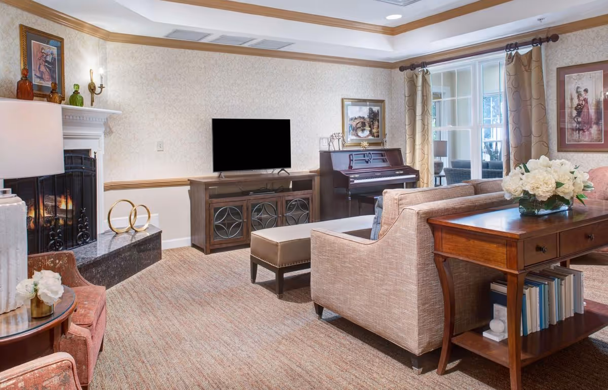 A cozy living room with a beige sofa, a wooden console table with white flowers in a vase, a piano, a TV on a wooden stand, a fireplace with a fire burning, and framed artwork on the walls. The room has carpeted flooring and large windows with curtains.