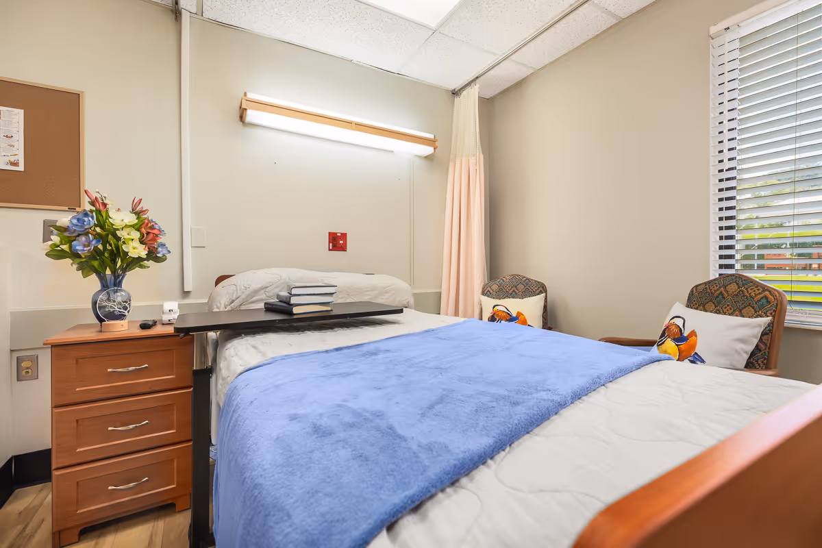 A neatly made hospital-style bed with a blue blanket and white sheets in a healthcare facility room. There is a wooden bedside table with a vase of colorful flowers and a few books on an overbed table. Two cushioned chairs with decorative pillows featuring bird designs are positioned near a window with white blinds. The walls are beige, and there is a fluorescent light fixture above the bed.