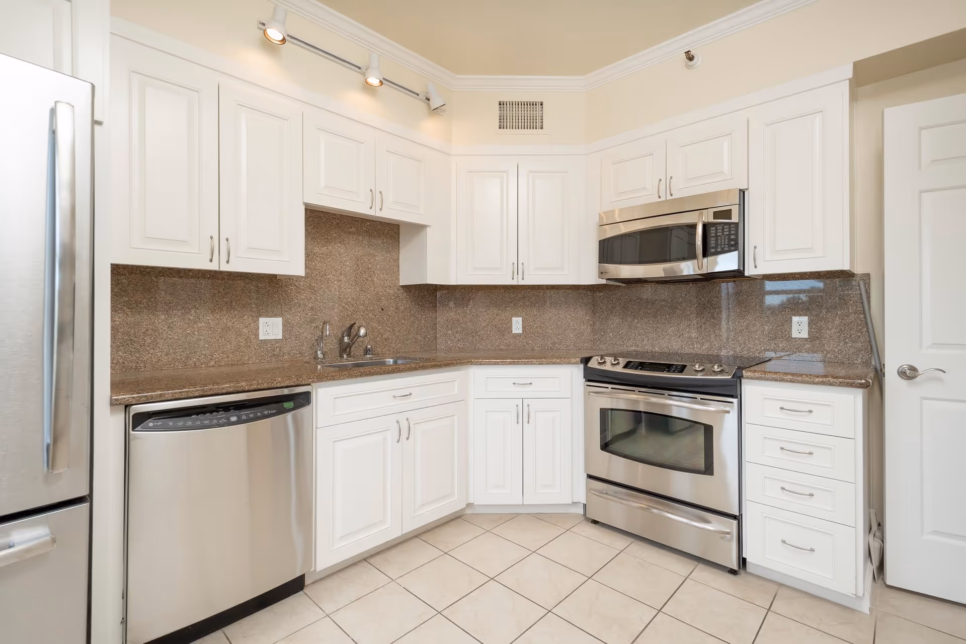 Modern kitchen with white cabinets, stainless steel refrigerator, dishwasher, oven, and microwave. The countertops and backsplash are brown granite, and the floor is tiled in a light color.