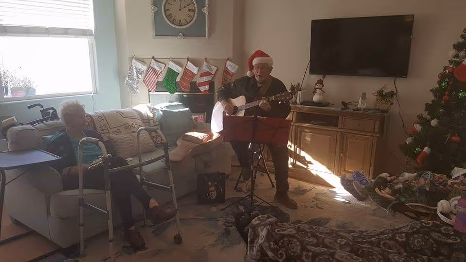 A cozy living room decorated for Christmas with a Christmas tree, stockings hanging on the wall, and holiday decorations. An elderly man wearing a Santa hat is playing a guitar and singing, while an elderly woman sits on a couch nearby with a walker in front of her. The room has a TV mounted on the wall and a wooden cabinet beneath it.