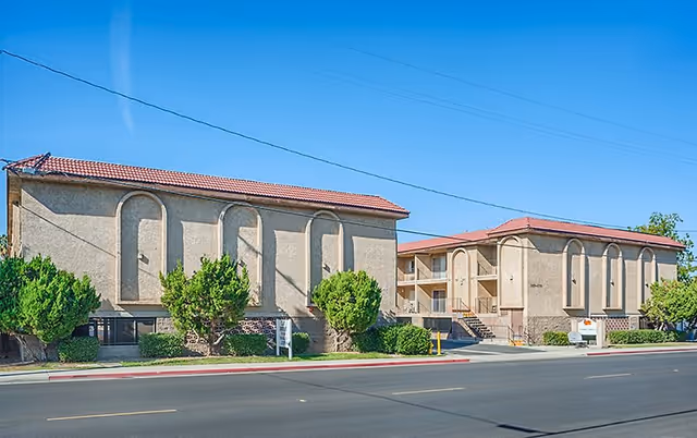 Exterior view of Vista Montana Senior Living facility showing two beige stucco buildings with red tile roofs, small trees and bushes in front, and a clear blue sky above.