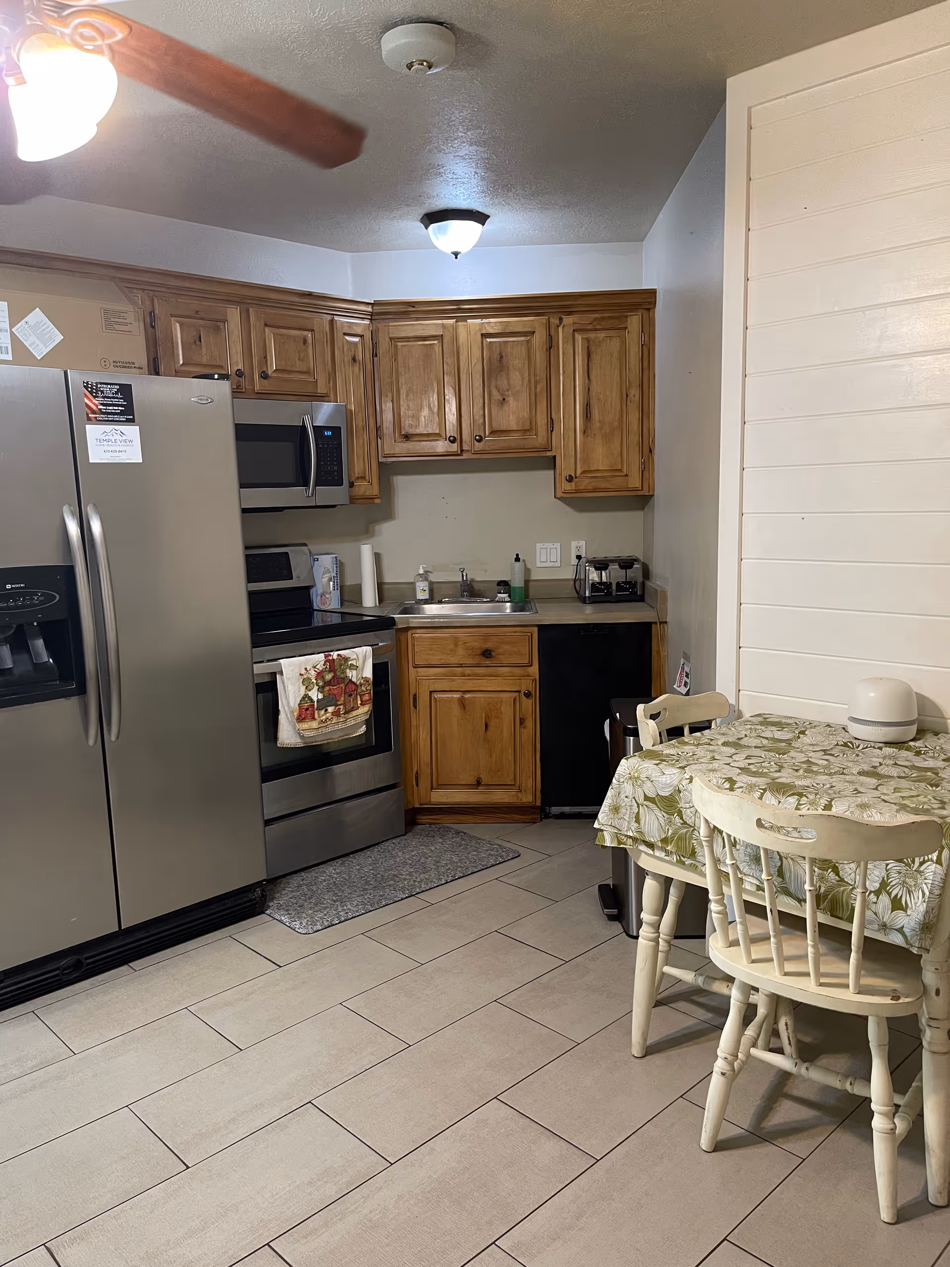 A kitchen area with wooden cabinets, a stainless steel refrigerator, microwave, and stove. There is a small dining table with a floral tablecloth and two white wooden chairs. The floor is tiled, and there is a ceiling fan with a light fixture visible.