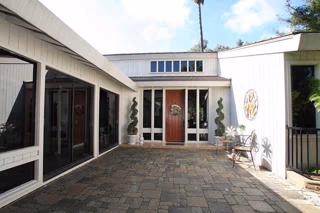 Outdoor courtyard area of a care home with a paved stone floor, white exterior walls, large windows, a wooden front door with a wreath, two spiral-shaped topiary plants in pots, a small table with two chairs, and a decorative circular wall art piece.