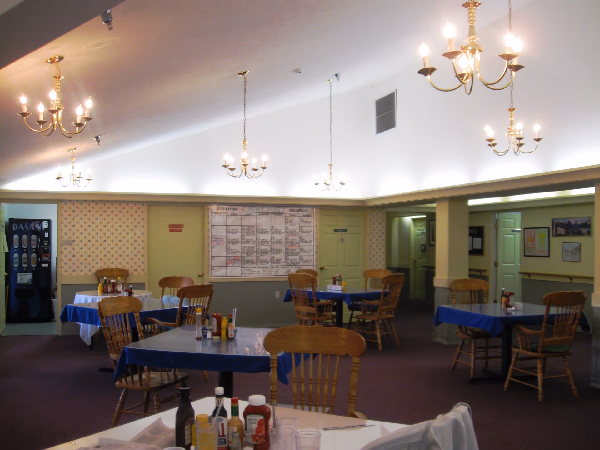 Interior view of a dining room with several wooden tables and chairs. Tables are covered with blue tablecloths and have condiments like ketchup, mustard, and hot sauce. The room has multiple brass chandeliers hanging from the ceiling, wallpaper with a floral pattern, and a large activity calendar on the far wall. There is a vending machine in the back left corner.