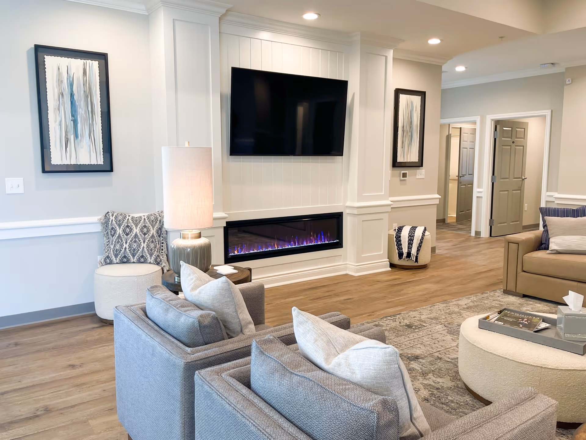 Bright modern living room with gray armchairs and sofa, a wall-mounted TV above an electric fireplace, and neutral decor.