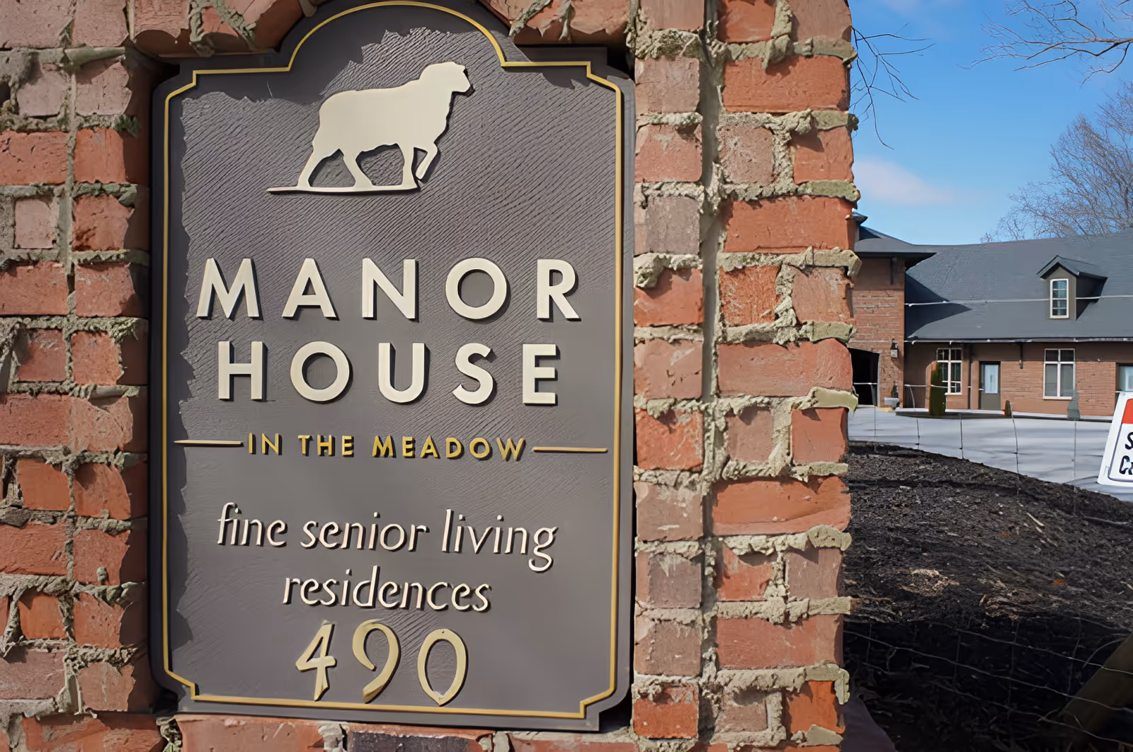 Decorative brick sign reading "Manor House in the Meadow: fine senior living residences 490" with the facility building visible in the background.