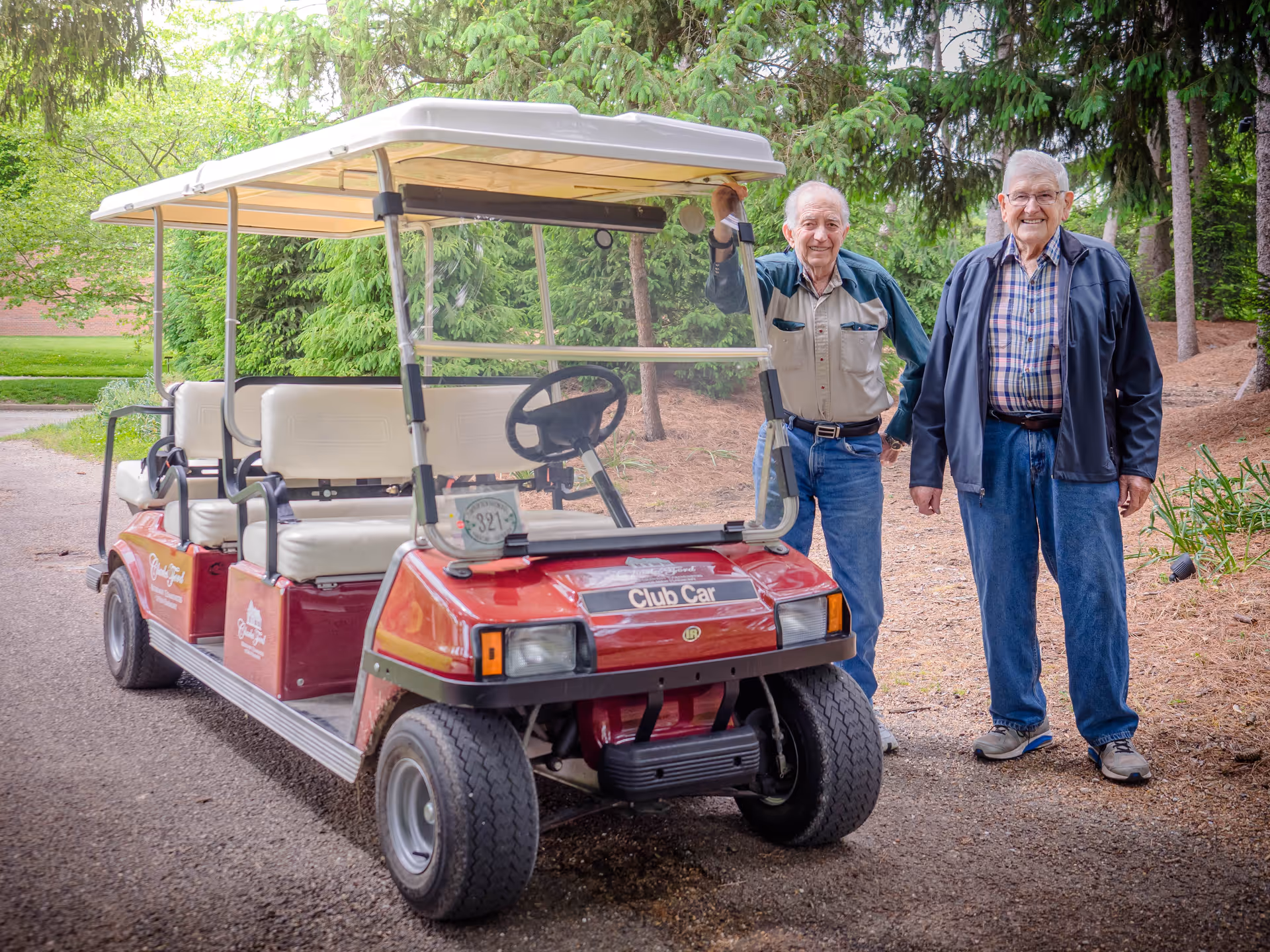Two elderly men standing outdoors next to a red Club Car golf cart on a paved path surrounded by trees and greenery.
