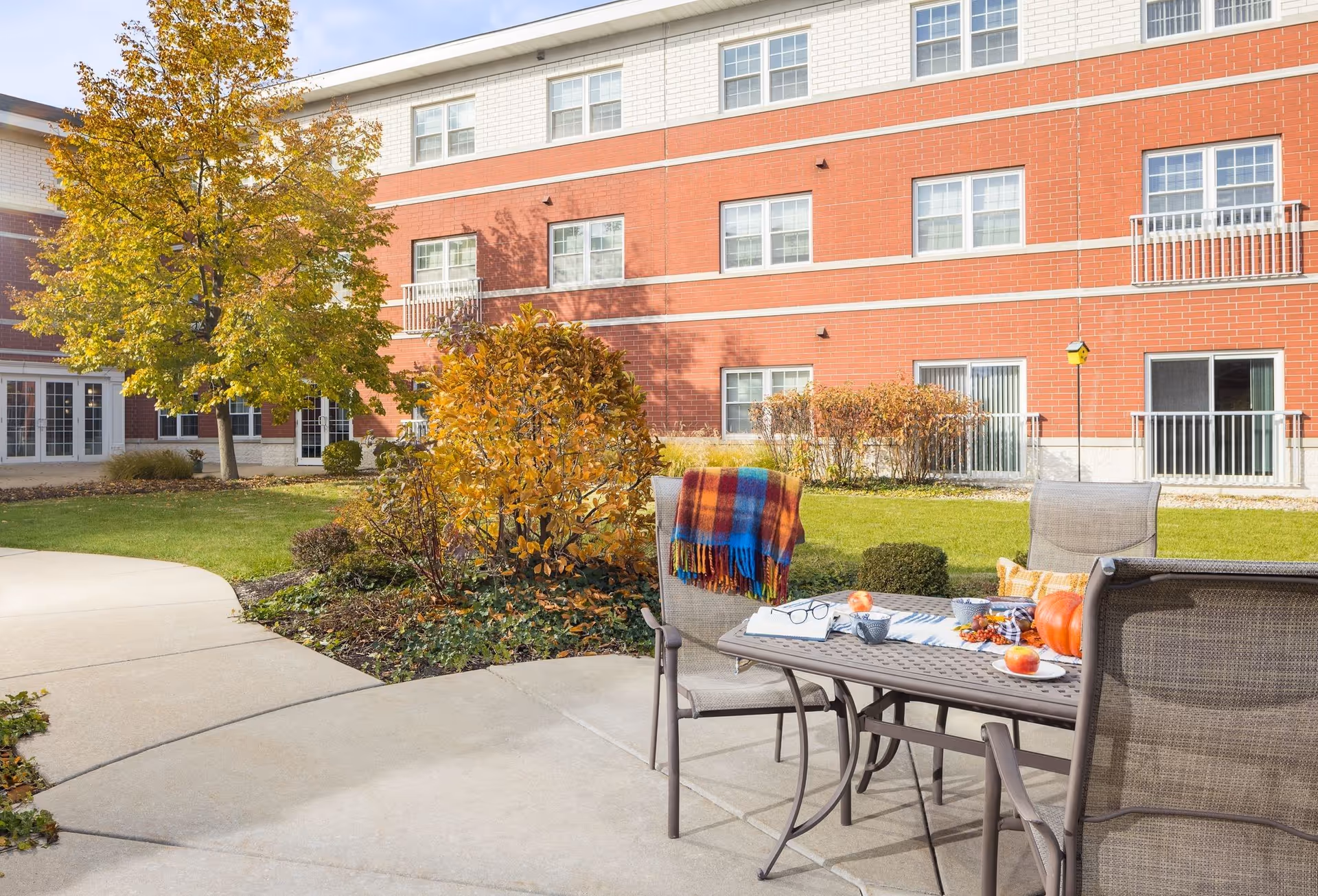 Outdoor patio area at Hanover Place with a table and four chairs. The table is decorated with a colorful blanket draped over one chair, a pair of glasses, a cup, a pumpkin, and some fruit. The patio is surrounded by green grass, bushes with autumn-colored leaves, and a tree with yellow leaves. The background shows a red brick building with multiple windows and sliding glass doors.