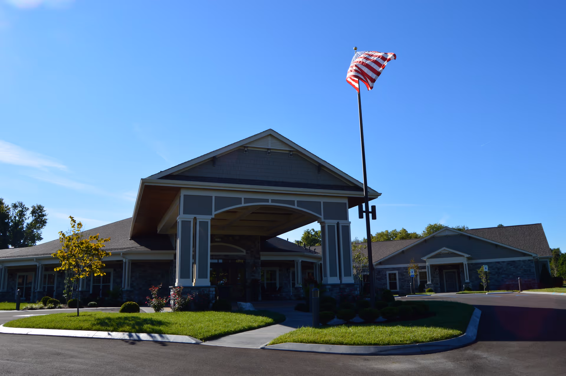 Exterior view of Morning Pointe of Louisville facility showing the main entrance with a covered driveway, manicured landscaping, and an American flag on a tall flagpole against a clear blue sky.