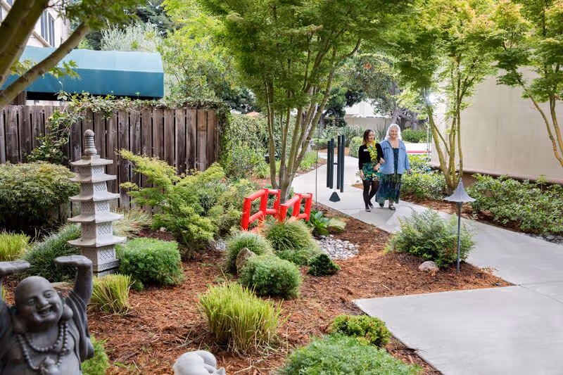 Two women walking along a paved pathway in a landscaped garden area with green trees, shrubs, and decorative elements including a small pagoda statue and a laughing Buddha statue.