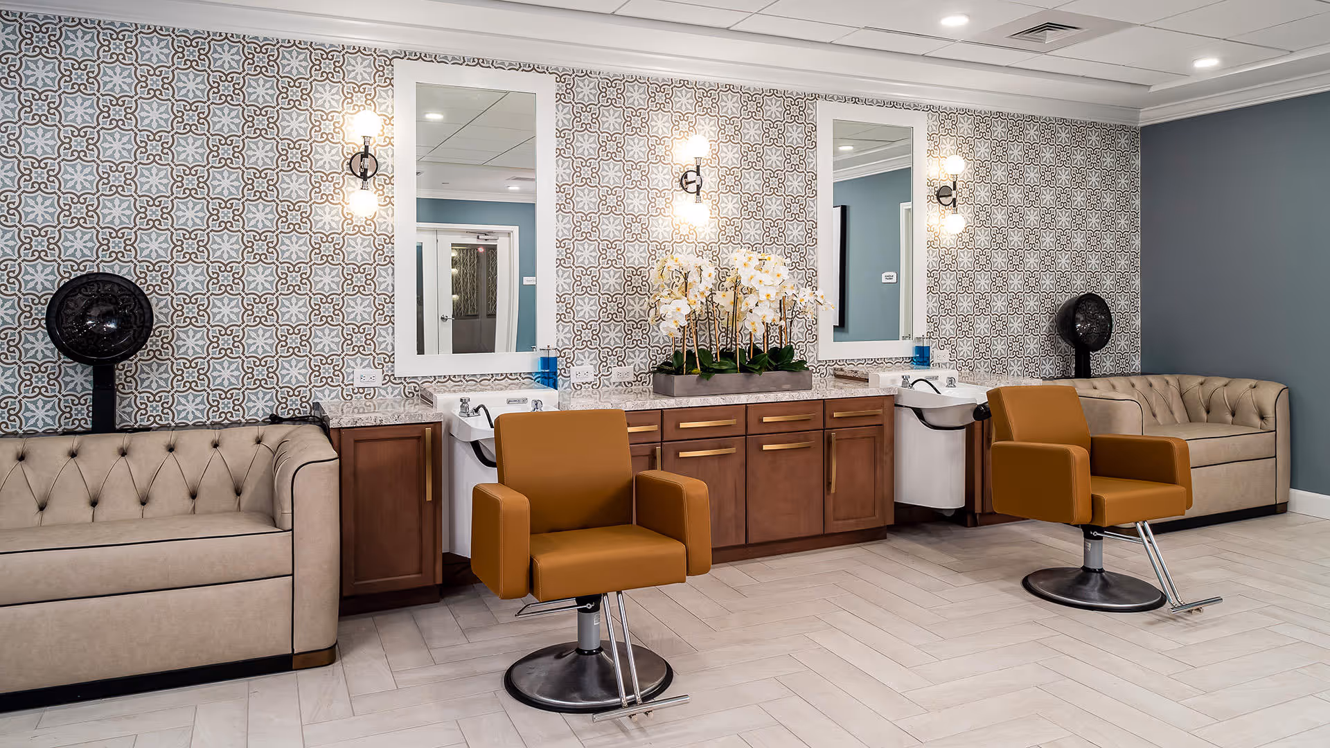 Interior of a salon area in a senior living facility with two brown salon chairs in front of sinks and mirrors. The wall behind features patterned wallpaper and two beige tufted sofas on either side. There are wall-mounted hair dryers and a decorative arrangement of white orchids on the countertop.