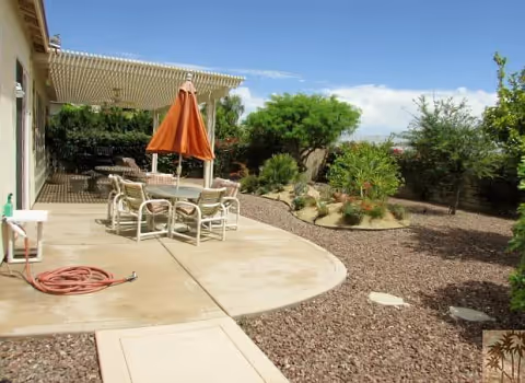 Backyard patio with a dining table and chairs under a pergola and orange umbrella beside a gravel landscaped yard with shrubs and trees.