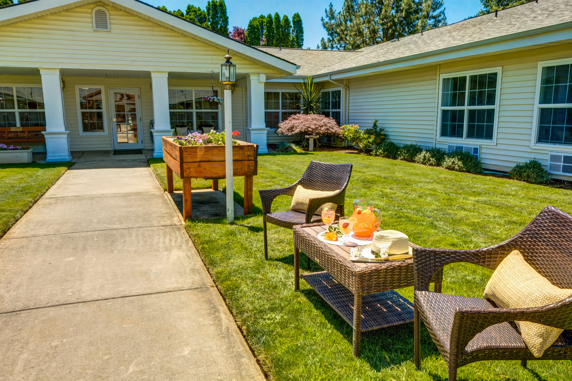 Outdoor seating area at Morrow Heights Assisted Living with two wicker chairs and a wicker table on a green lawn. The table has a pitcher of orange drink, two glasses, a hat, and some snacks. The building with white siding and multiple windows is in the background, along with a concrete walkway leading to the entrance.