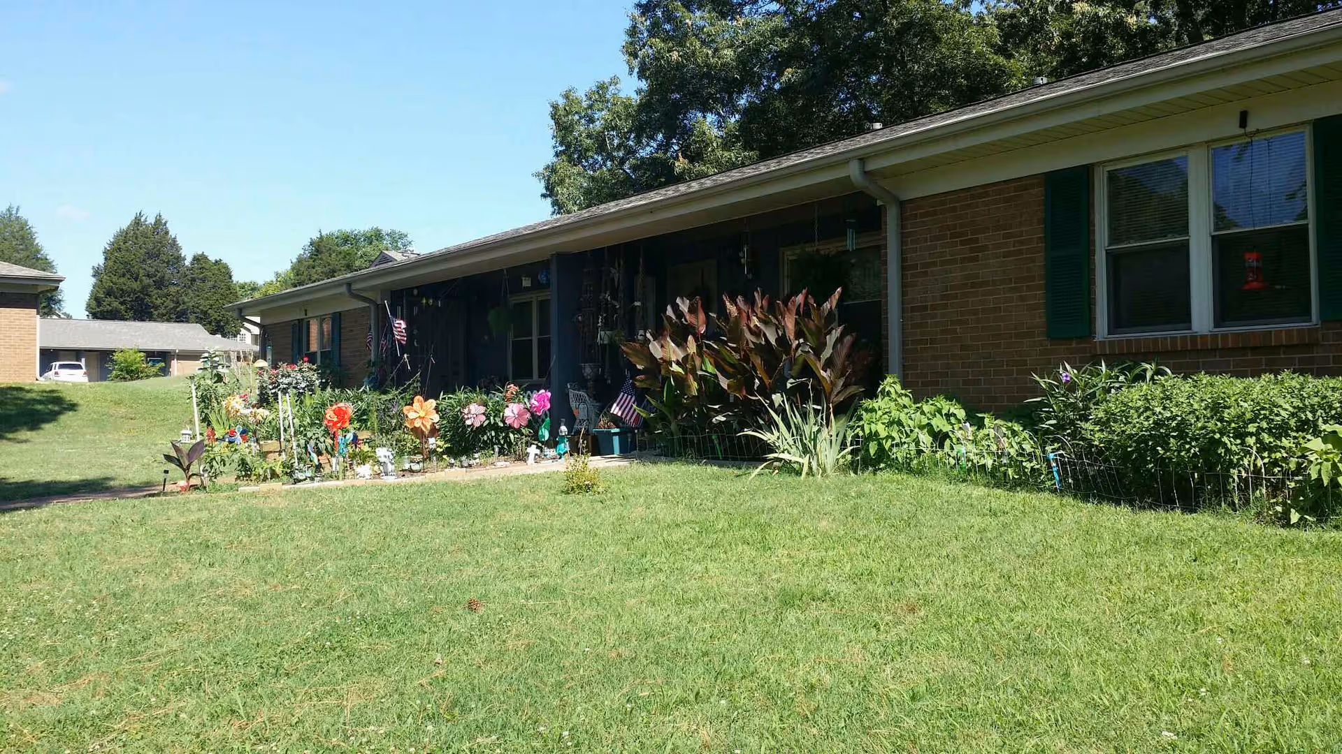 Exterior view of a single-story brick building with green shutters and a well-maintained lawn. There is a garden bed with various colorful flowers and plants in front of the building. The sky is clear and blue, and there are trees in the background.
