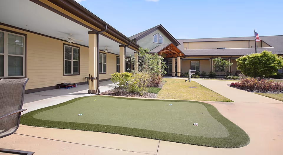 Outdoor courtyard area at Rosecastle at Deerwood featuring a putting green with golf balls, surrounded by a paved walkway, garden beds with shrubs, and a covered porch with ceiling fans attached to the building. The building has beige siding and a peaked roof with an American flag visible in the background under a clear blue sky.